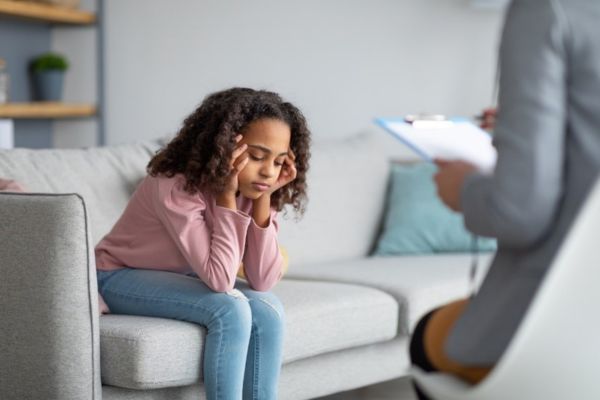 little girl sitting on  a couch with hands on face looking sad speaking to a therapist 