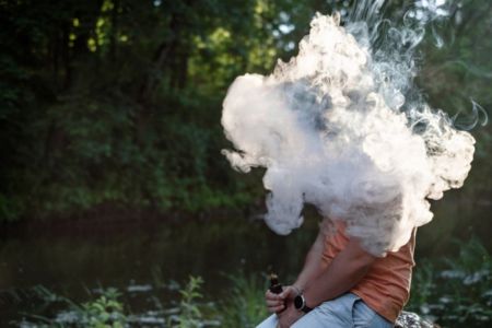 a young adult sitting outdoors wiht a vape in hand and surrounding by vape smoke